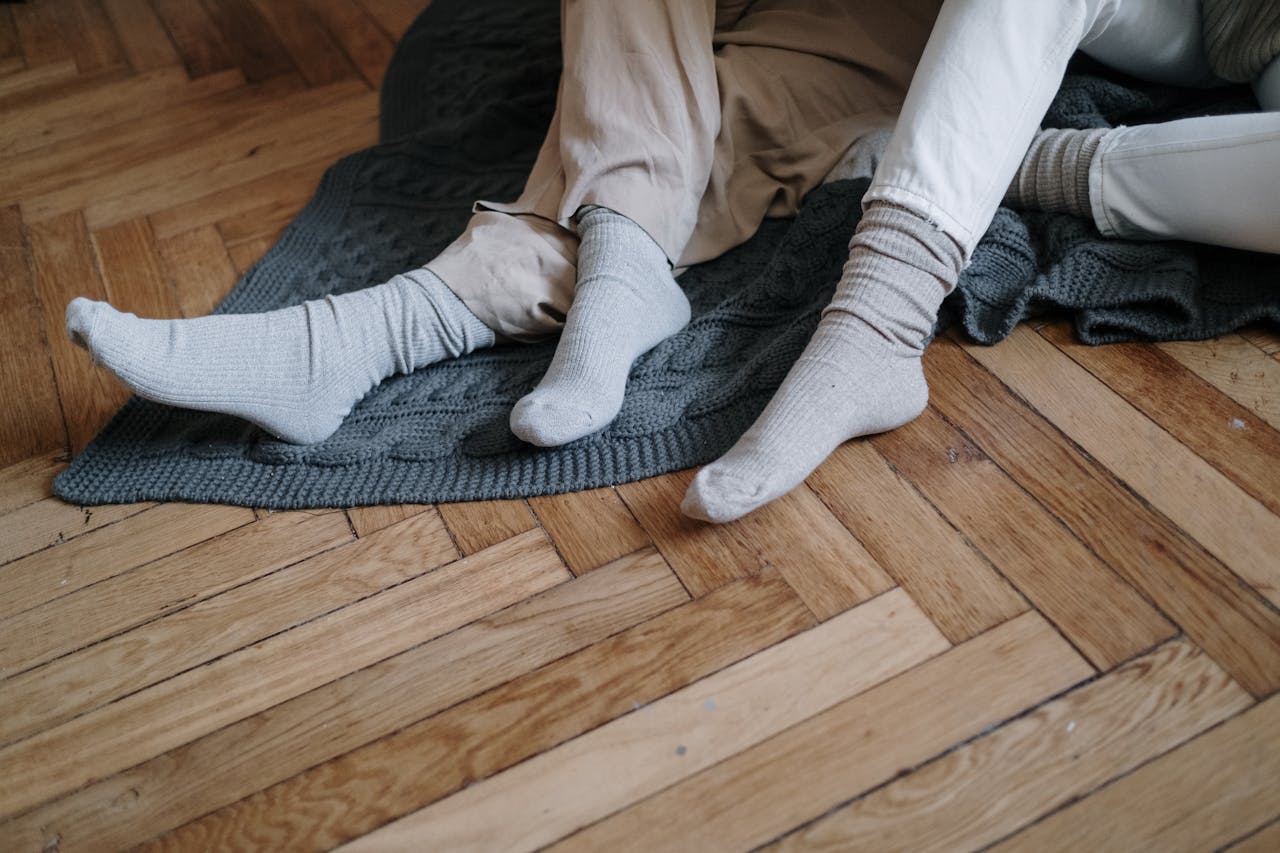 A couple wearing socks, relaxing on a wooden floor with a gray blanket, enjoying warmth.
